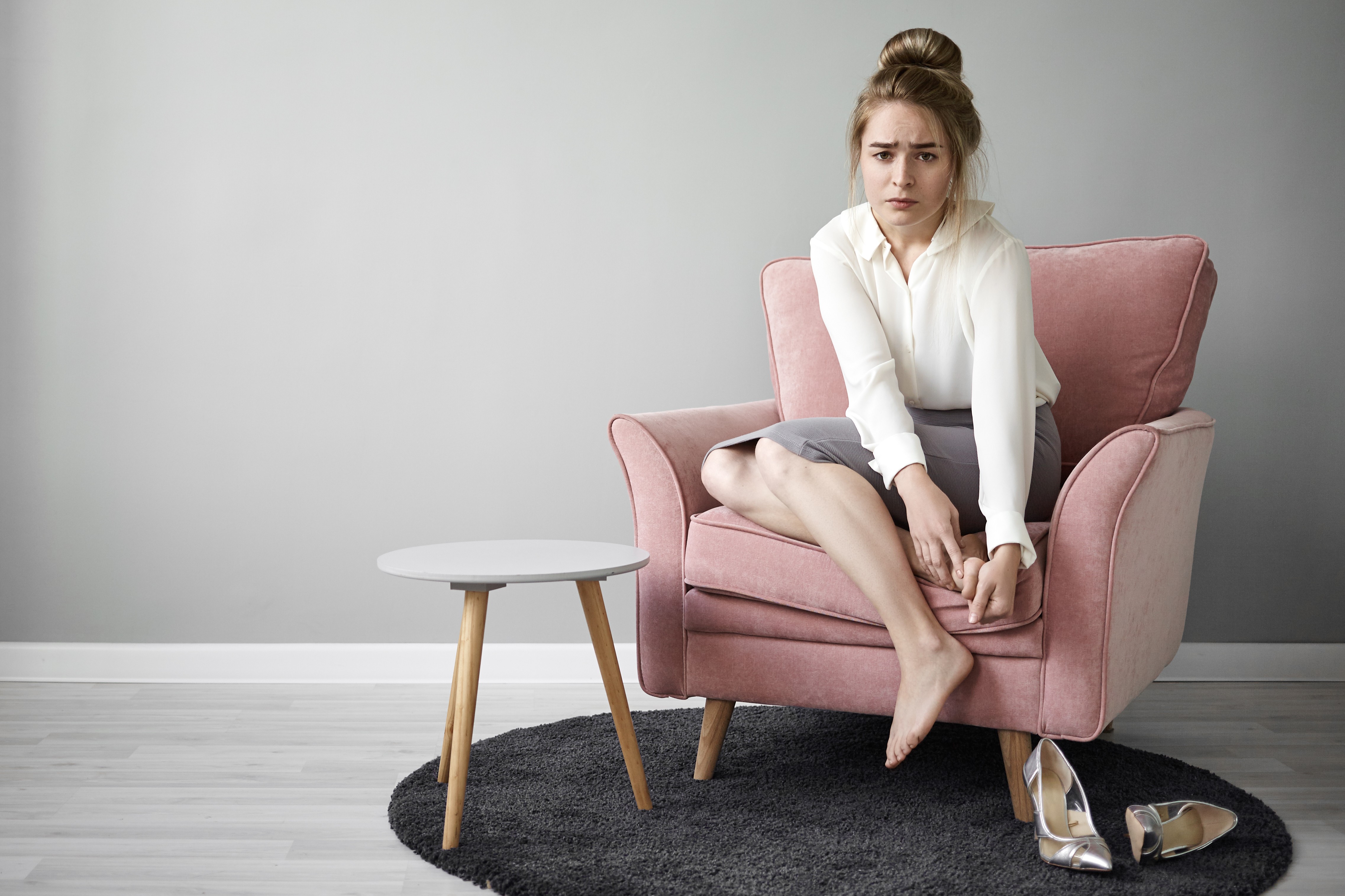 Mulher jovem, com coque alto e expressão de desconforto, sentada descalça em poltrona rosa, massageando os pés. Veste blusa branca e saia cinza. Sapatos prateados estão no chão, sobre tapete escuro, ao lado de uma mesa lateral branca.