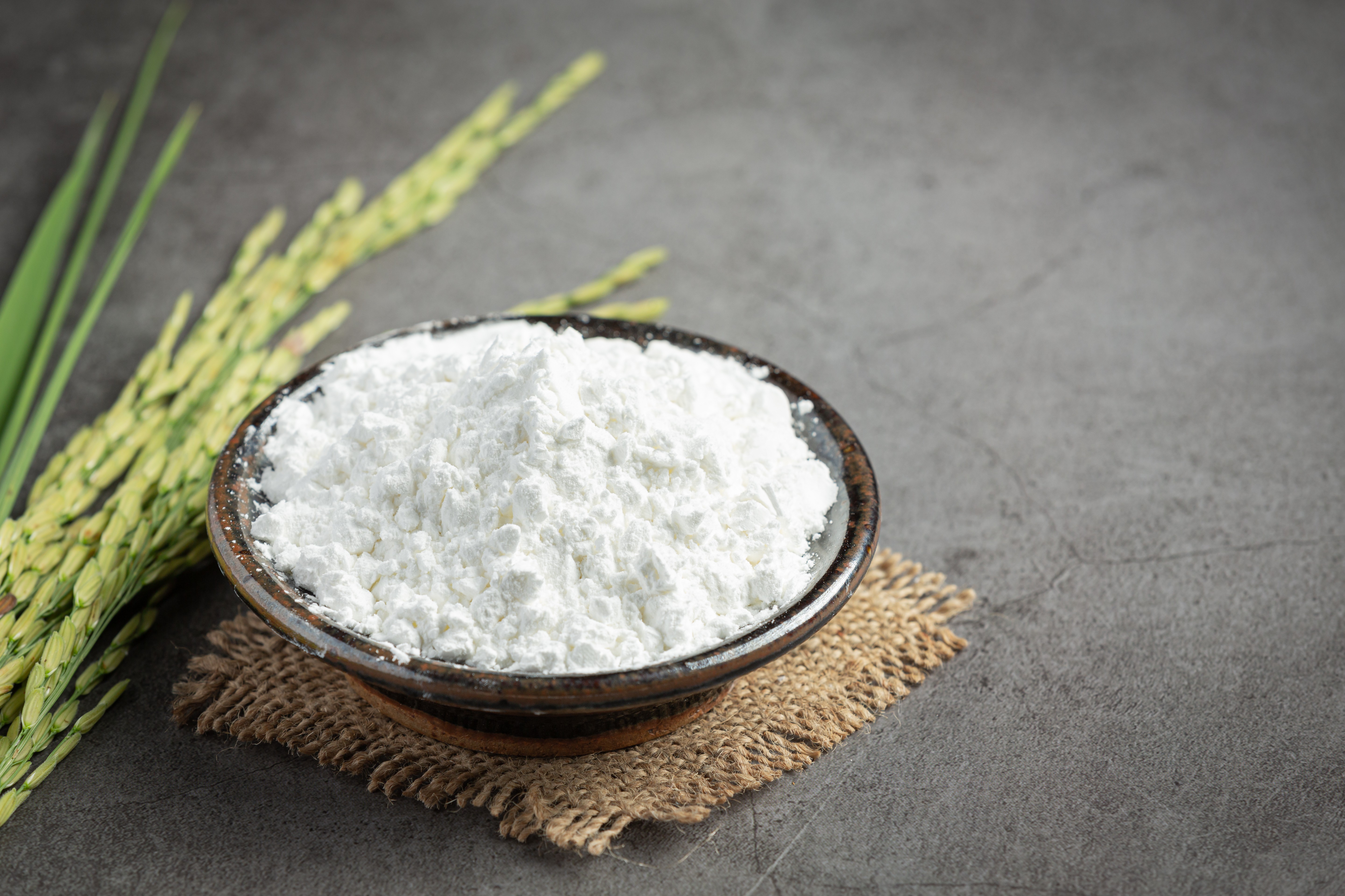 Tigela de cerâmica escura cheia de farinha branca sobre um pedaço de juta, com espigas de arroz verde à esquerda, em um fundo cinza escuro