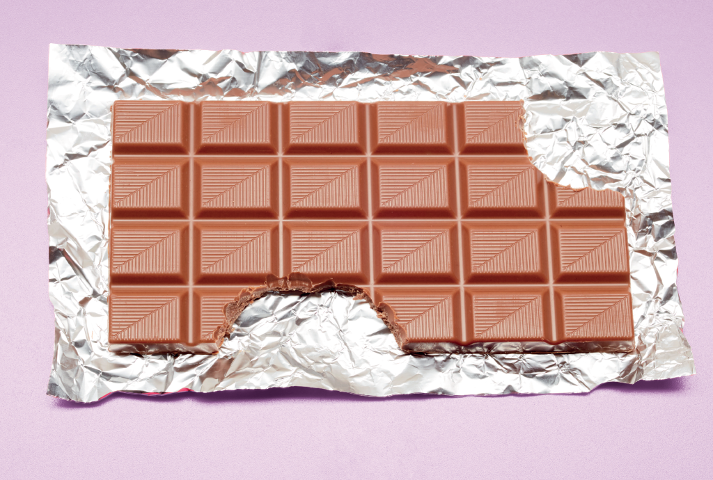 Barra de chocolate ao leite parcialmente comida, com uma mordida na parte inferior, sobre papel alumínio amassado e fundo lilás