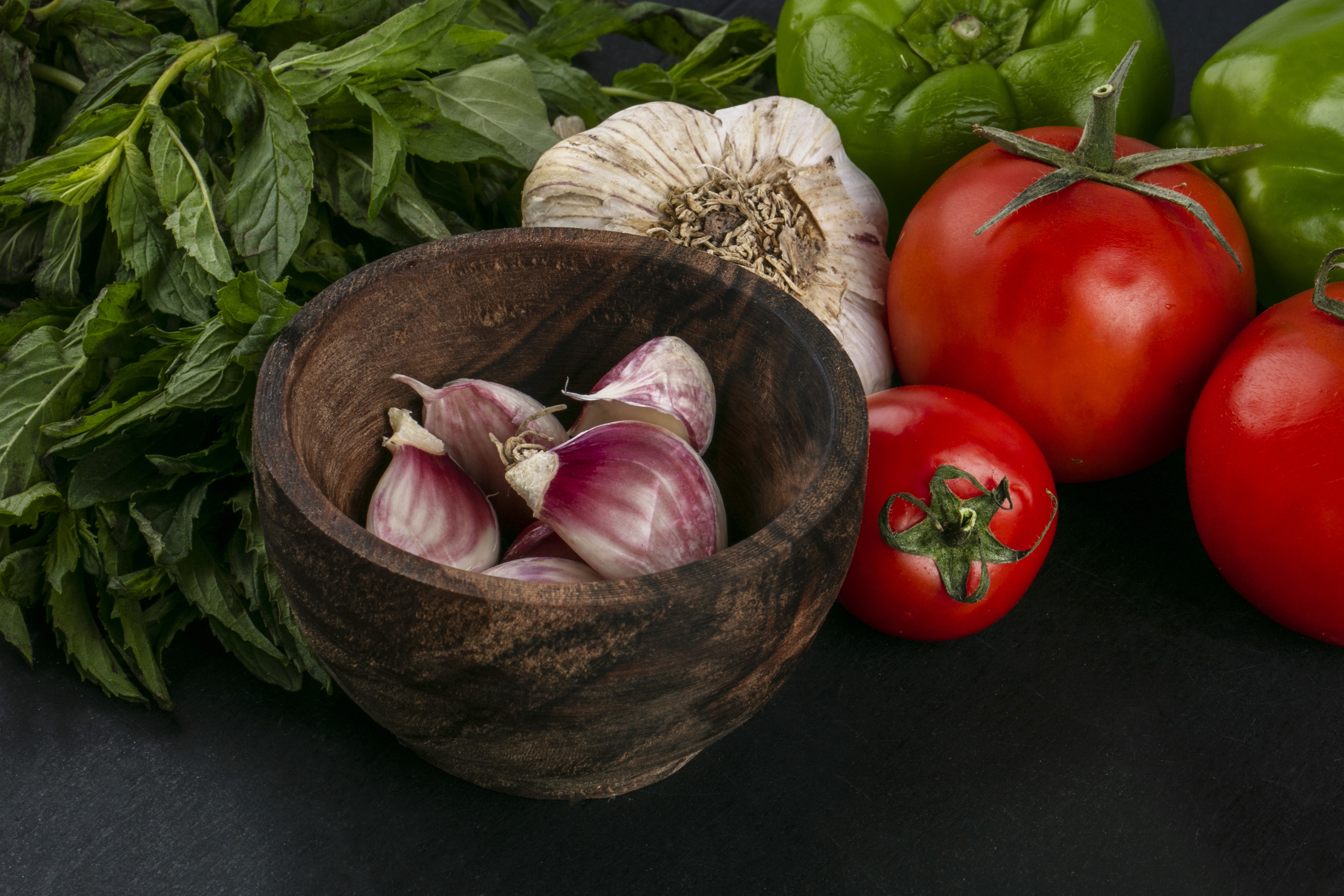 Alho descascado em tigela de madeira, cabeça de alho, pimentões verdes, tomates vermelhos e folhas de hortelã fresca sobre fundo escuro