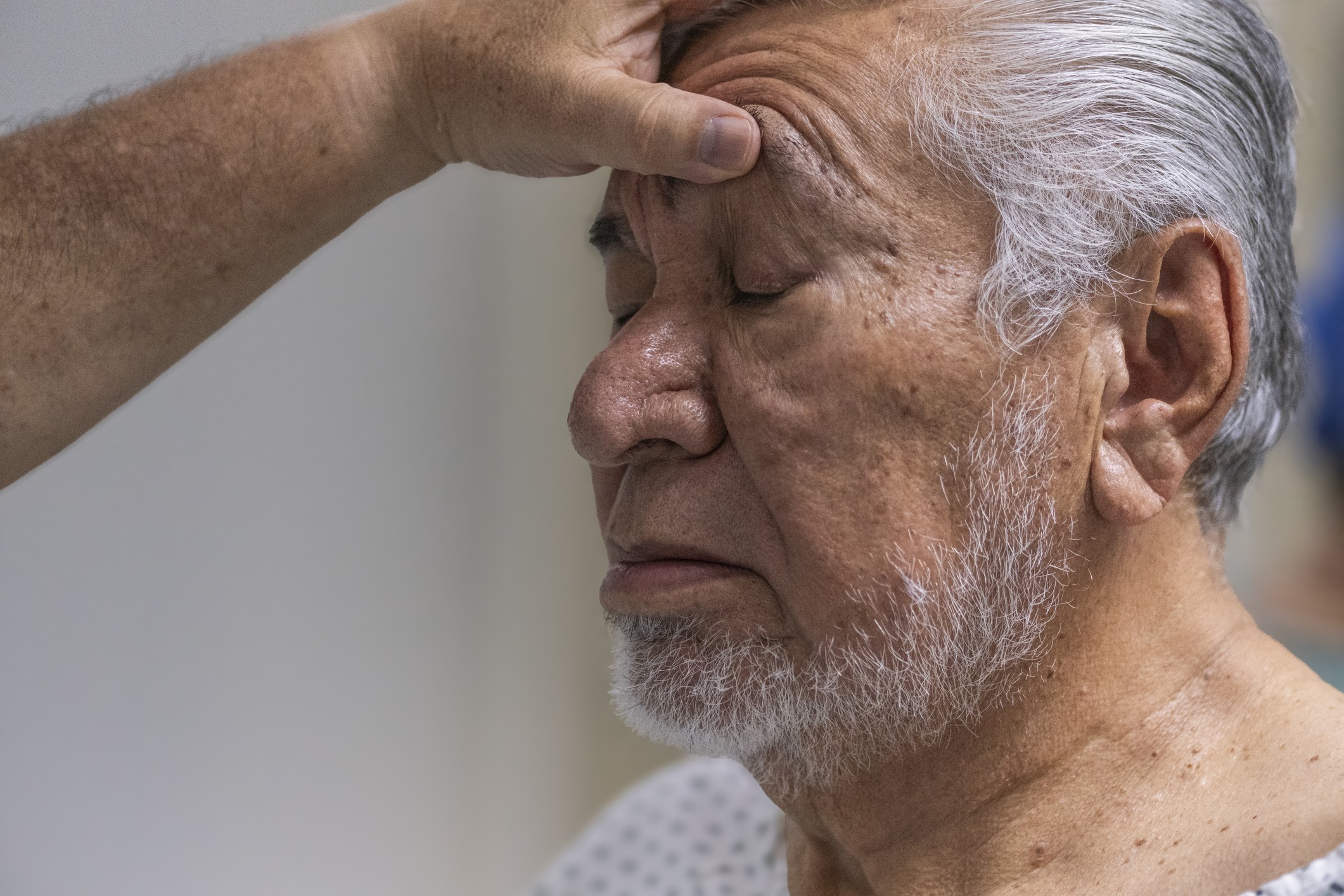 Homem idoso de pele morena e cabelos grisalhos, com barba branca, olhos fechados e expressão serena, enquanto uma mão toca sua testa. Ele veste uma camisa branca com bolinhas cinzas.
