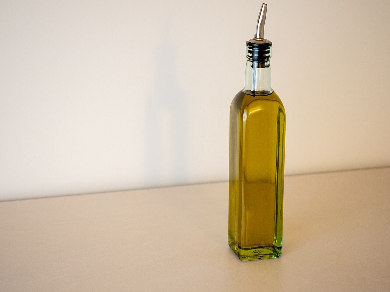 Garrafa de azeite de oliva em vidro verde-claro com bico dosador metálico, sobre uma superfície bege e fundo branco com sombra