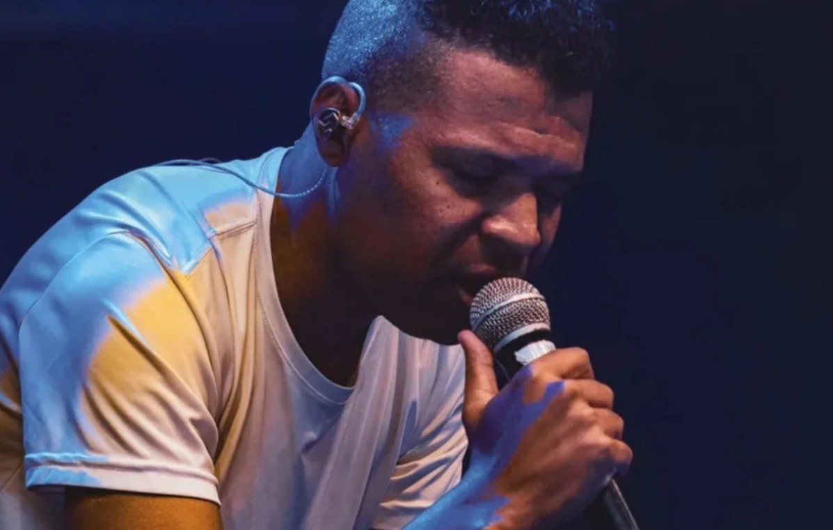 Homem negro, cabelo curto e crespo, usando fones de ouvido intra-auriculares, canta com os olhos fechados segurando um microfone. Veste camiseta branca com reflexos azuis e amarelos da iluminação do palco.