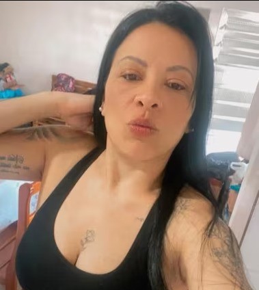 Mulher de cabelos pretos longos, com tatuagens nos bra&ccedil;os e no peito, vestindo top preto, fazendo bico para a c&acirc;mera em um ambiente interno