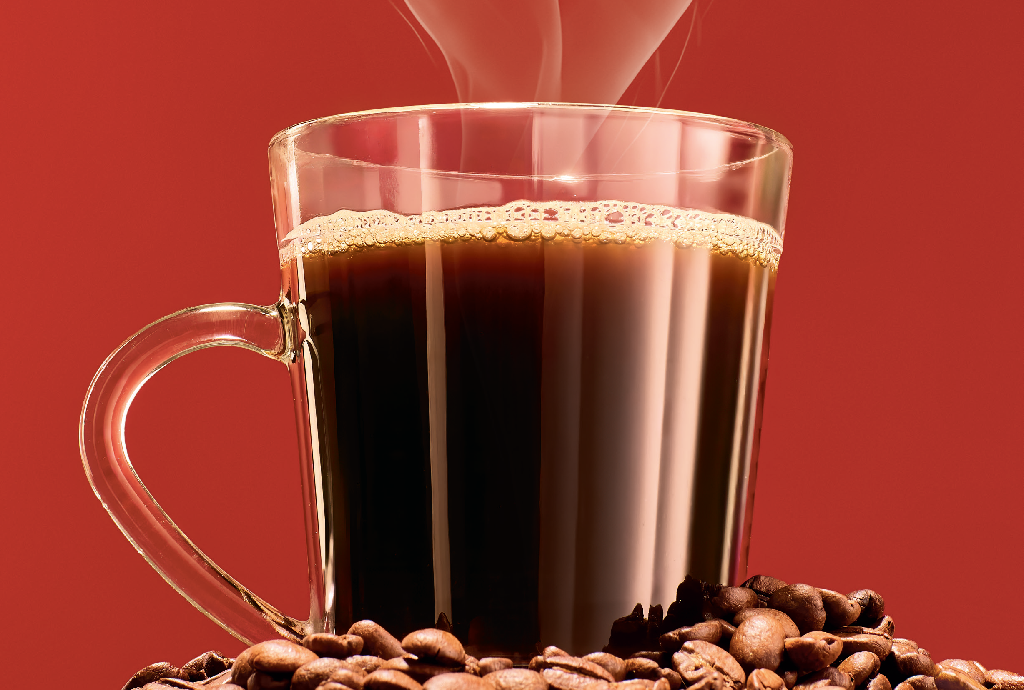 Caneca de vidro transparente com café preto fumegante e espuma, cercada por grãos de café torrados, sobre fundo vermelho