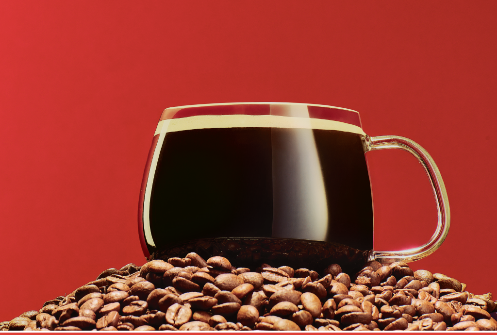 Xícara de vidro transparente com café preto sobre uma pilha de grãos de café torrados, em fundo vermelho