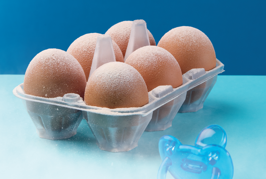 Seis ovos marrons em uma embalagem plástica transparente, com uma fina camada de pó branco sobre eles. Ao lado, uma chupeta azul translúcida. O fundo é azul