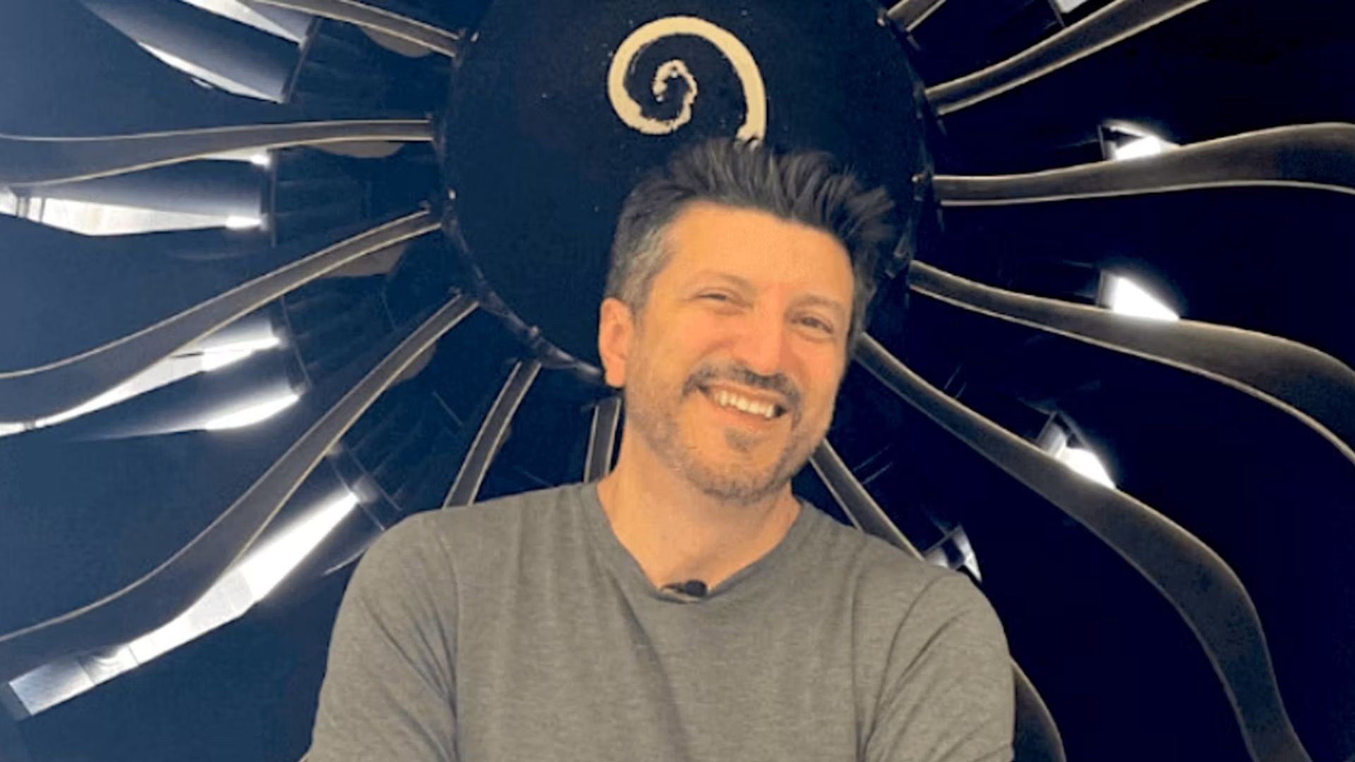 Homem de meia-idade, cabelo escuro e barba rala, sorri para a câmera, vestindo camiseta cinza. Ao fundo, um motor de avião com hélices escuras e um espiral branco no centro