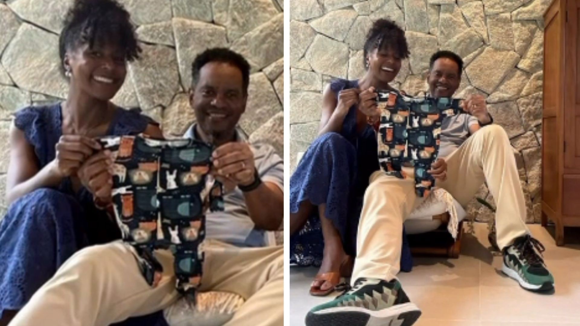 Um casal sorridente, mulher de vestido azul e homem de camisa polo clara e calça bege, segura um macacão de bebê estampado com gatos, em frente a uma parede de pedras