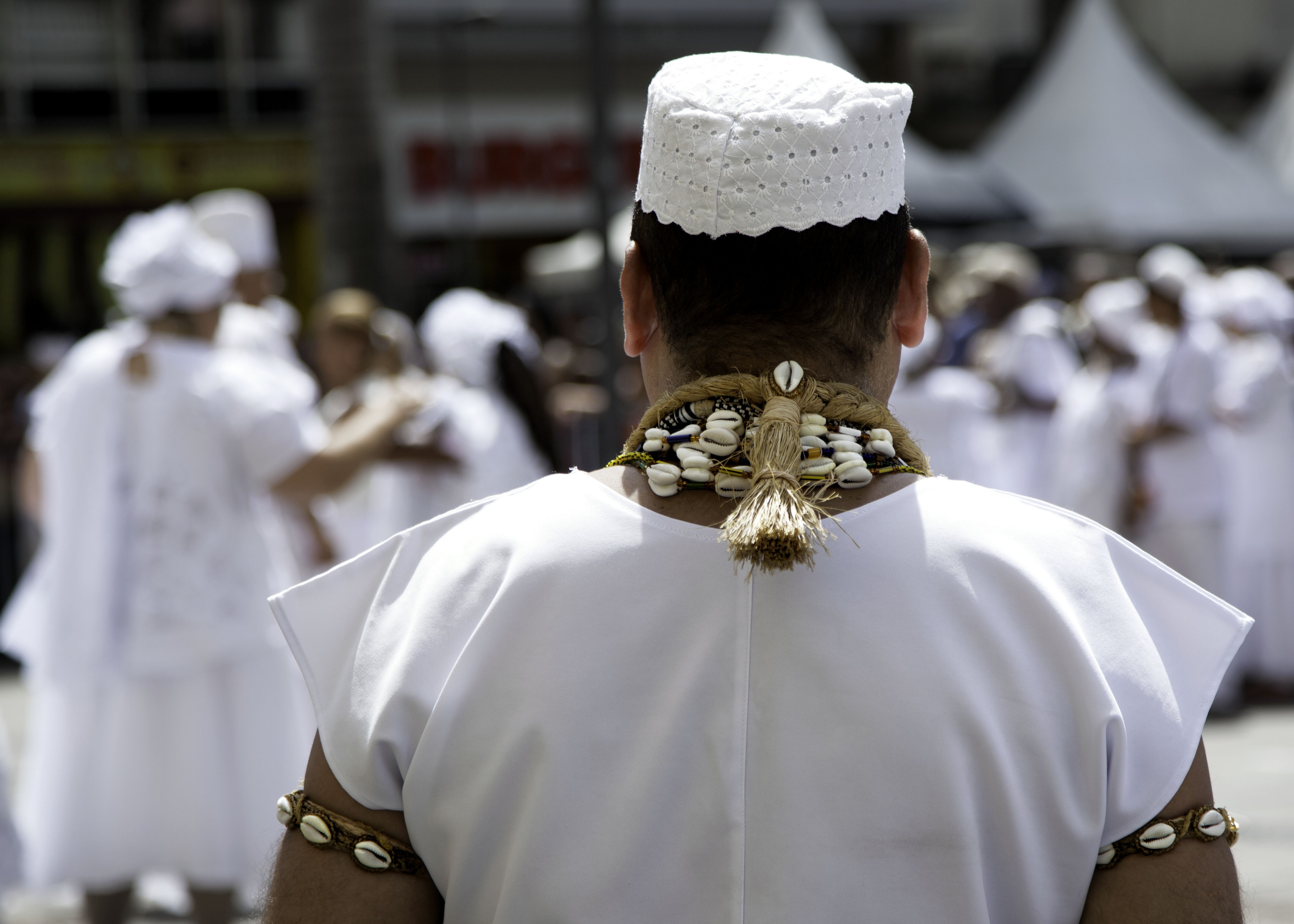 Homem de costas, vestindo túnica branca e gorro branco rendado, com colar e pulseira de búzios e palha. Ao fundo, pessoas borradas em roupas brancas e tendas brancas, sob luz solar