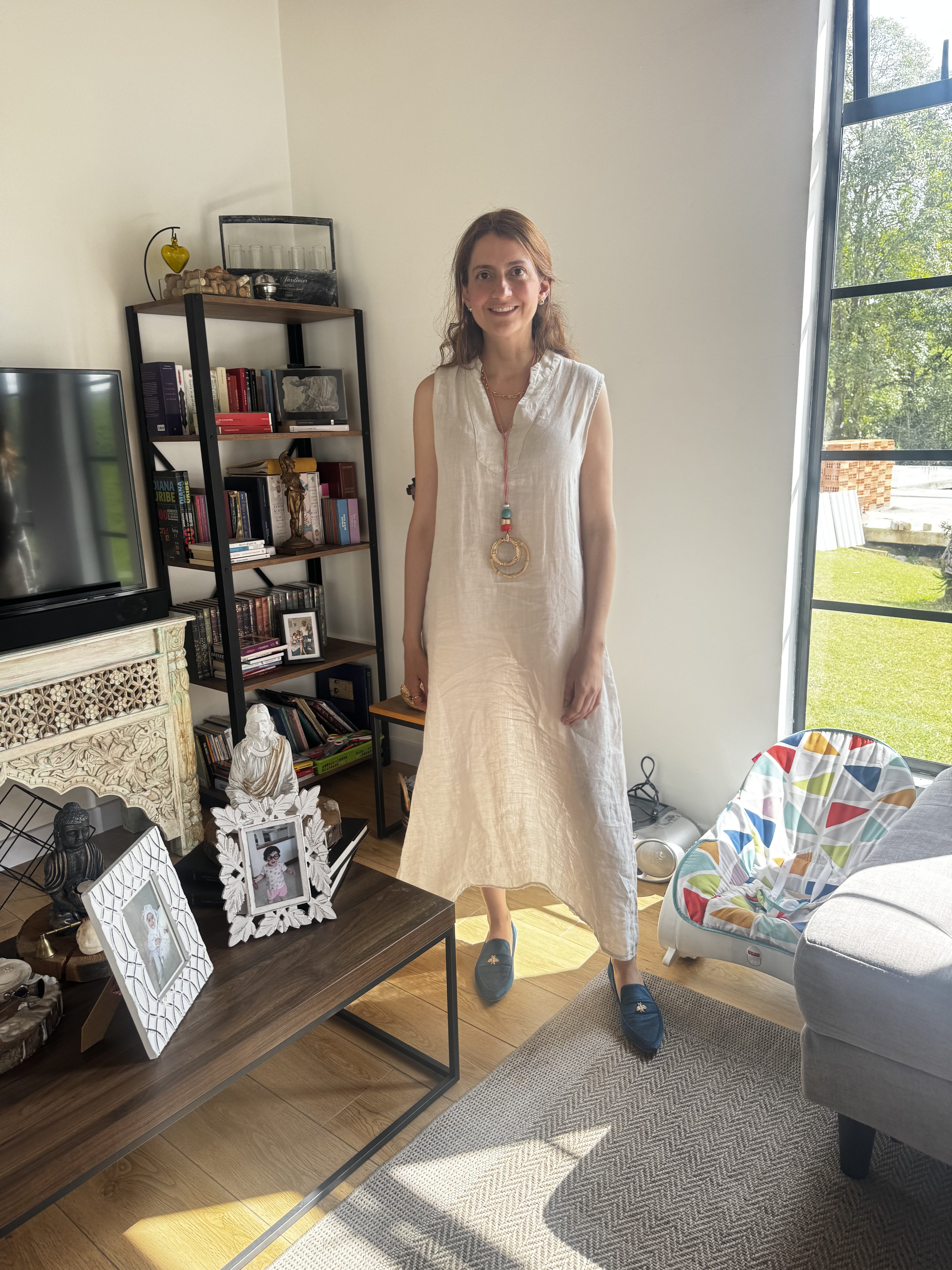 Mulher sorridente, de cabelos castanhos e vestido bege claro, em p&eacute; na sala de estar. Ela usa um colar com pingente circular e mi&ccedil;angas coloridas. Ao fundo, uma estante com livros e objetos decorativos, uma TV e uma janela grande com vista para o jardim. No ch&atilde;o, um tapete cinza e um ber&ccedil;o colorido