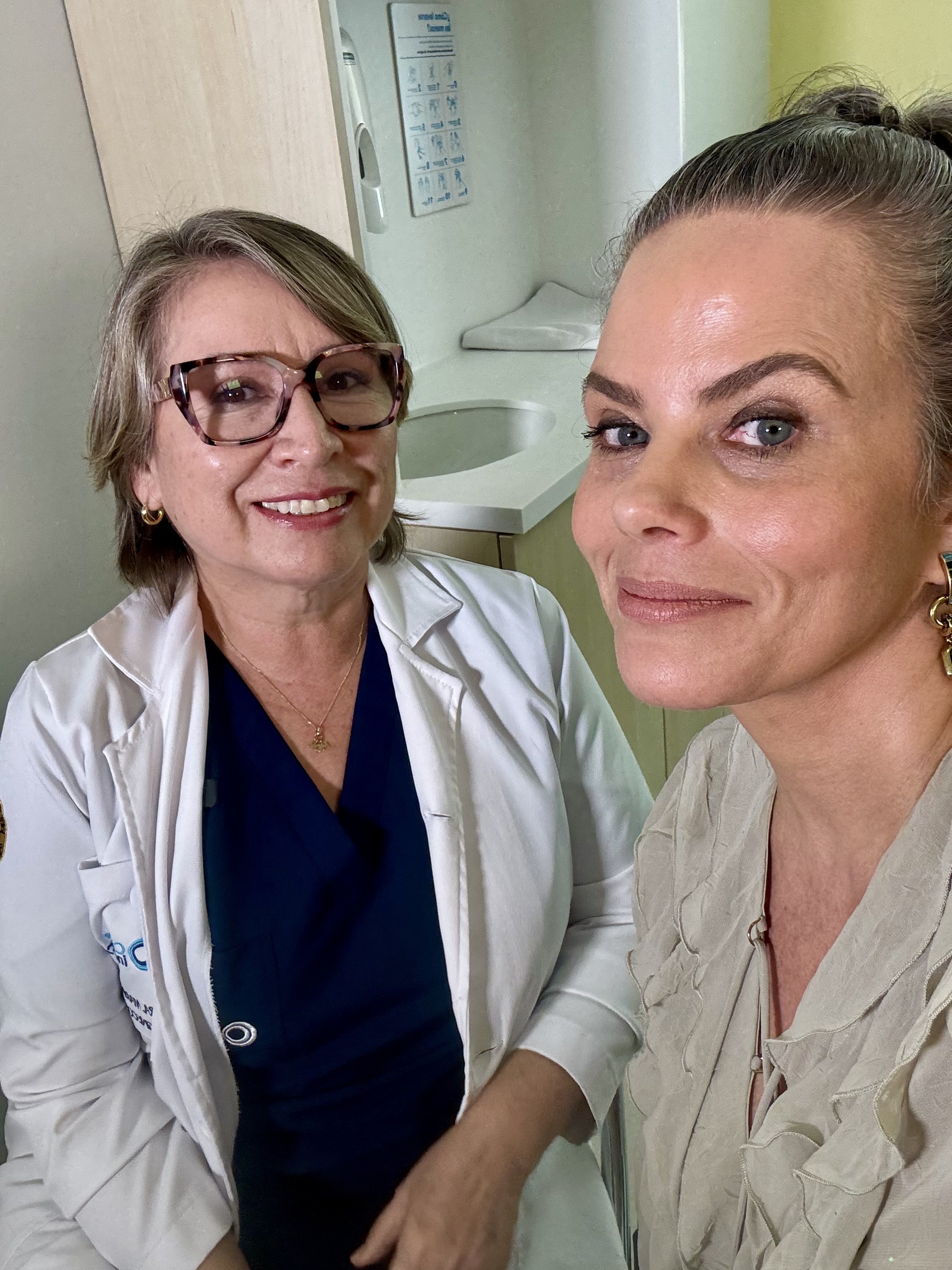 Duas mulheres sorrindo para a c&acirc;mera em um ambiente m&eacute;dico. &Agrave; esquerda, uma m&eacute;dica de jaleco branco e &oacute;culos, com um colar dourado. &Agrave; direita, uma mulher com blusa bege e brincos dourados, cabelo preso. Ao fundo, uma pia e um dispenser de papel.