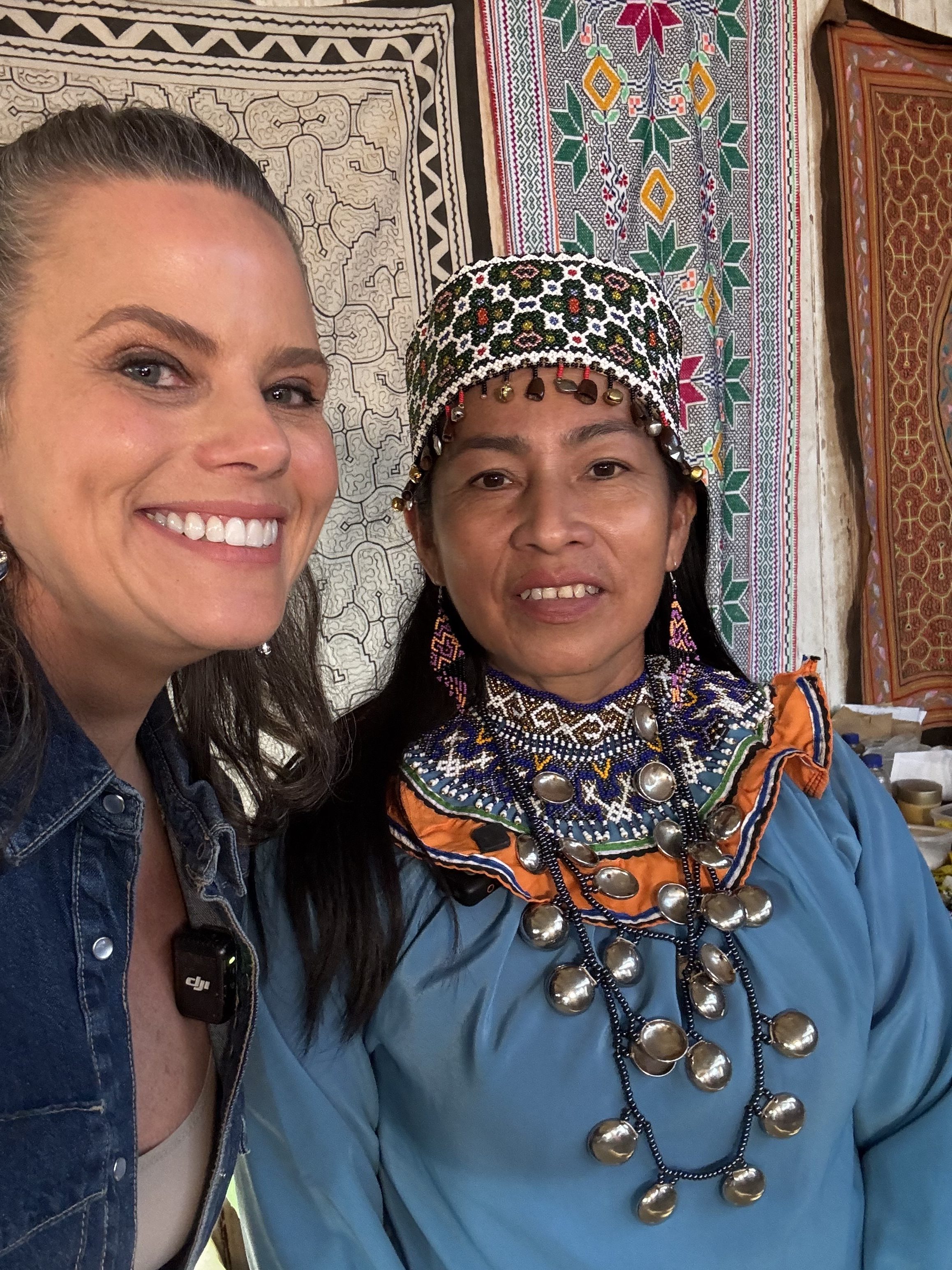 Duas mulheres sorrindo para a c&acirc;mera, uma com chap&eacute;u bordado e roupa azul com colar de moedas, a outra com jaqueta jeans, sobre tapetes estampados