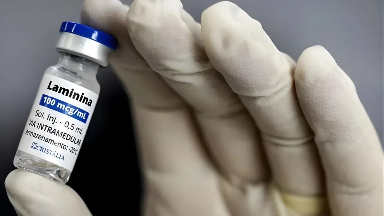Mão enluvada segurando um pequeno frasco de vidro com tampa azul e prata, contendo o medicamento Laminina 100 mcg/mL, solução injetável de 0,5 mL, para via intramedular, com armazenamento a -20°C, do laboratório Cristália