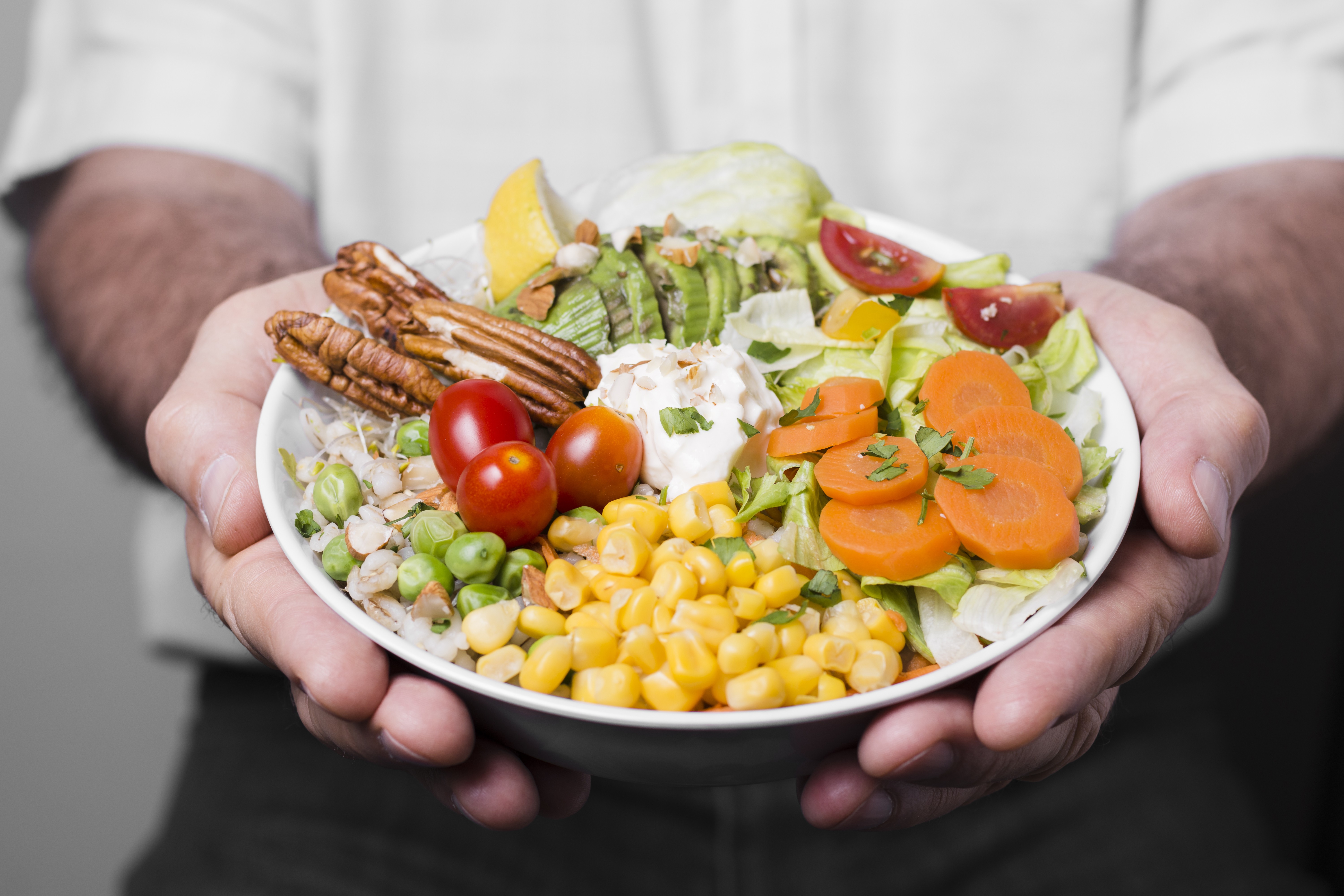 Homem de camisa branca segurando uma tigela de salada colorida com alface, milho, ervilhas, cenouras, tomates, abacate, nozes-pecã e um limão