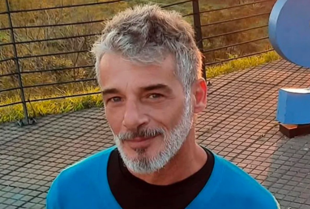 Homem de meia-idade com cabelo e barba grisalhos, vestindo camiseta azul sobre preta, sorri levemente para a câmera. Ele está em um ambiente externo, com grade e vegetação ao fundo, e um piso quadriculado.
