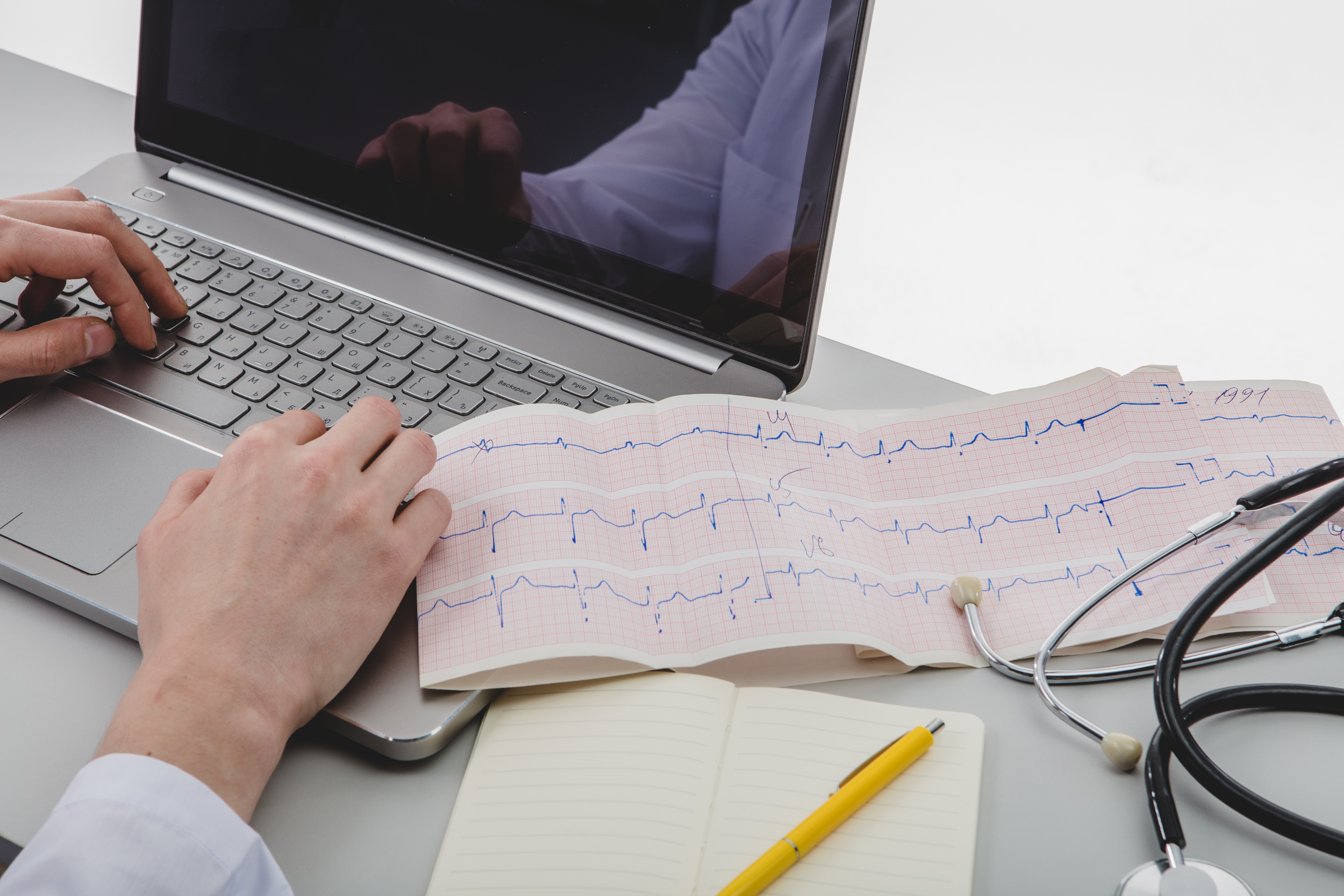 Mãos de um médico digitando em um laptop prateado, com um eletrocardiograma, um estetoscópio e um caderno com caneta amarela sobre a mesa
