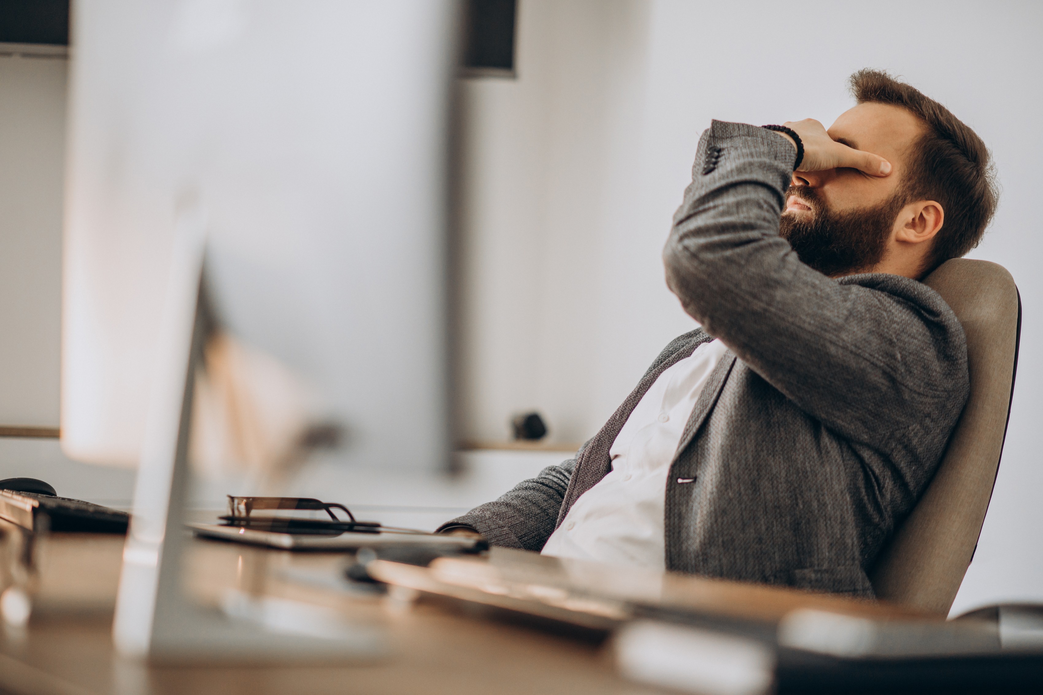 Homem barbudo de blazer cinza e camisa branca sentado em cadeira de escritório, com a mão direita cobrindo os olhos, parecendo cansado ou estressado, em frente a um monitor de computador em uma mesa de madeira