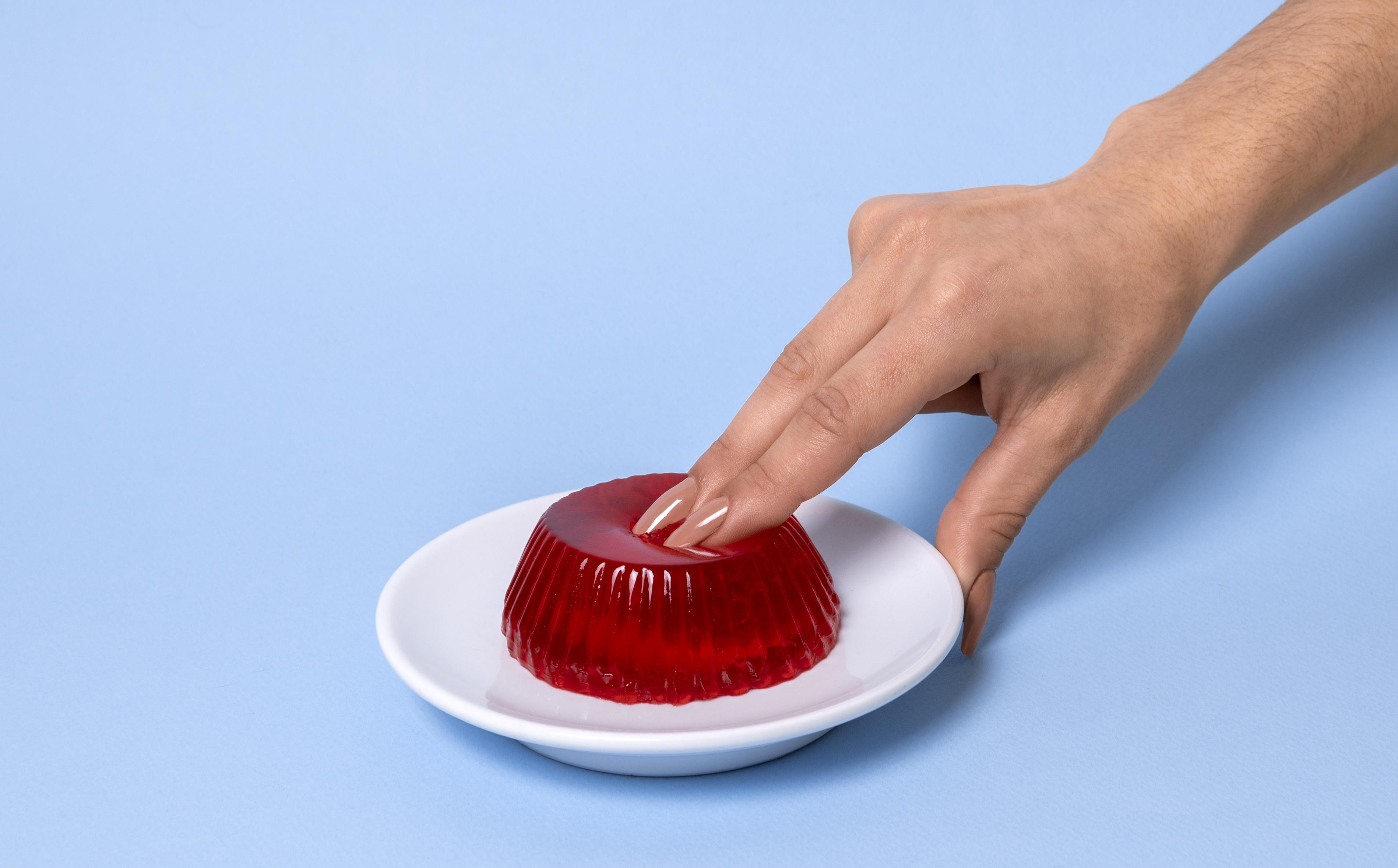 Mão feminina com unhas pintadas de nude pressionando levemente uma gelatina vermelha vibrante em um prato branco, sobre fundo azul claro