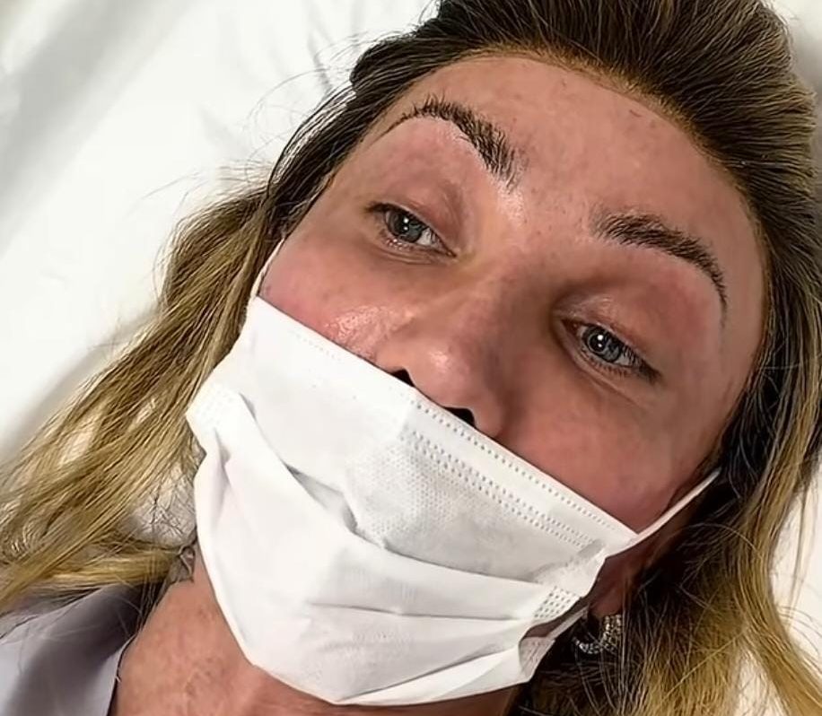 Mulher loira deitada, com máscara branca cobrindo nariz e boca, e sobrancelhas recém-desenhadas com pigmento escuro. Seu rosto e pescoço mostram vermelhidão e inchaço, indicando um procedimento estético recente. Ela veste uma blusa cinza e olha para cima com expressão séria