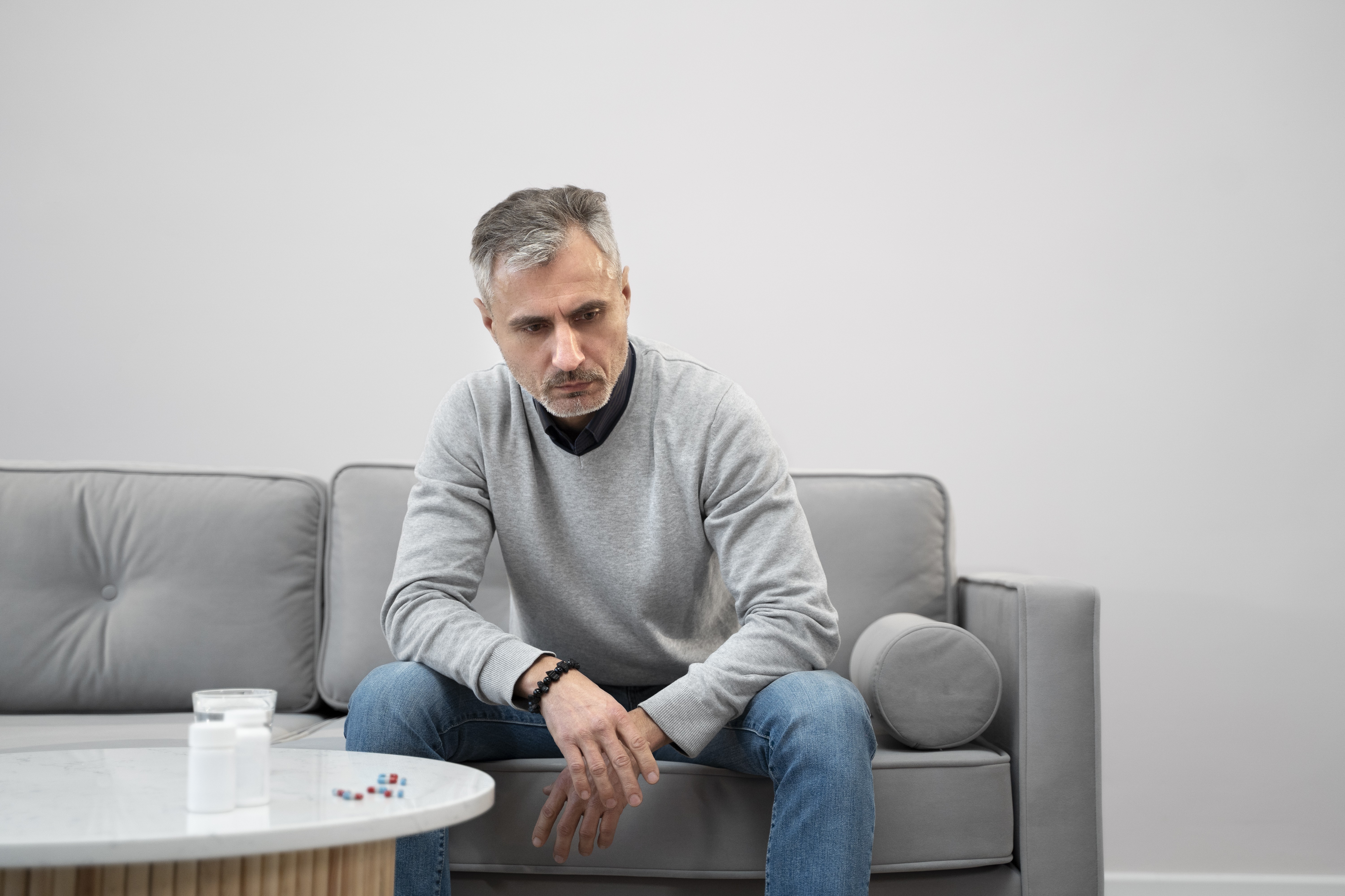 Homem de meia-idade, cabelos grisalhos, sentado em um sofá cinza, com expressão triste e olhar para baixo. Ele veste suéter cinza e calça jeans. Sobre a mesa de centro, há um copo dágua, dois frascos de remédio e algumas pílulas espalhadas