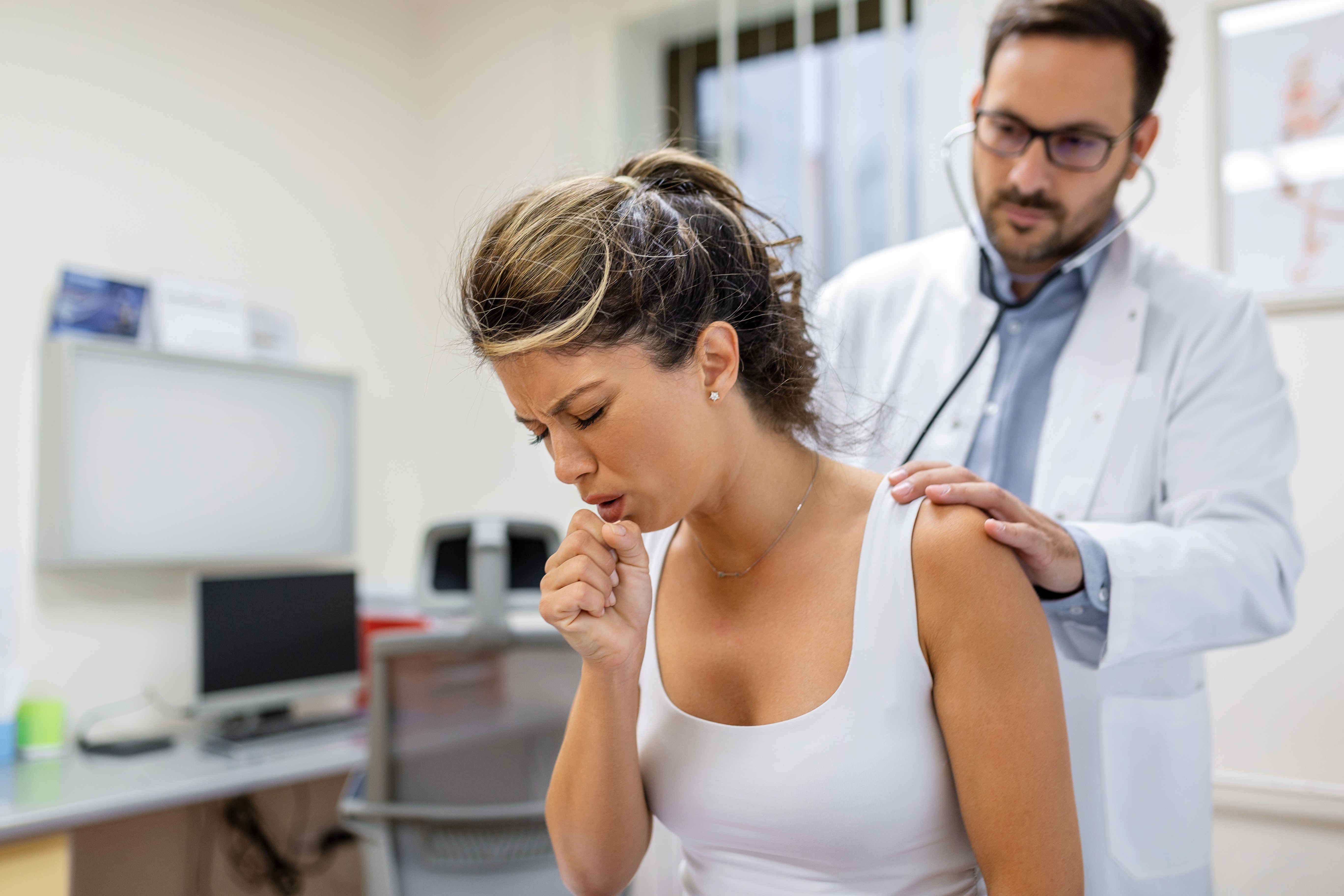 Mulher com cabelo preso, vestindo regata branca, tosse com a mão na boca enquanto um médico de jaleco branco e óculos a examina com estetoscópio e mão no ombro, em um consultório médico