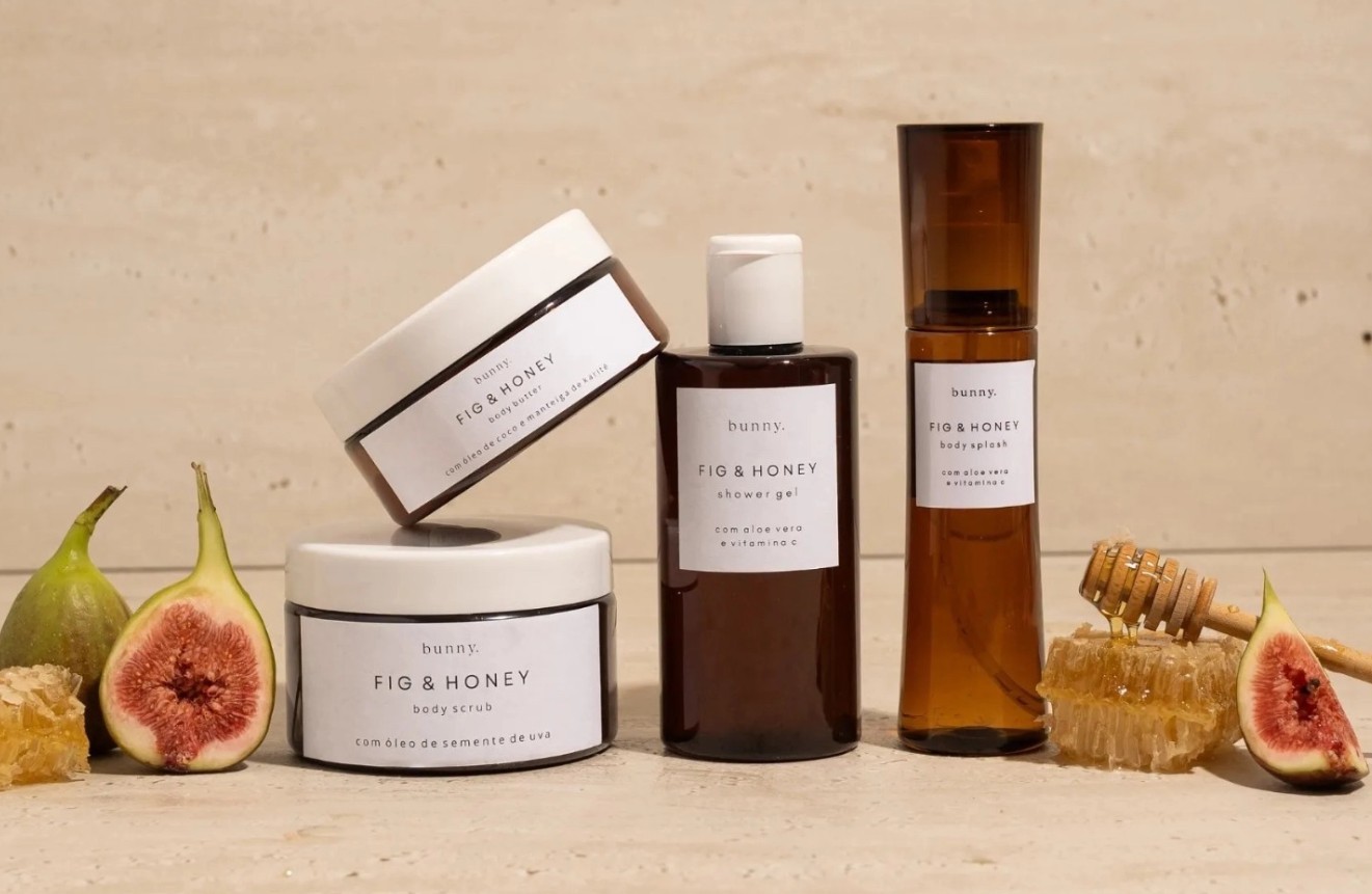 Produtos de beleza Bunny Fig & Honey: dois potes de creme, um frasco de gel de banho e um spray corporal, com figos frescos e favos de mel ao redor, sobre uma superfície clara
