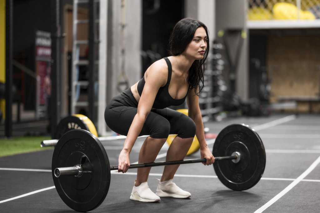 Mulher de cabelo escuro, vestindo top e cal&ccedil;a legging pretos, agachada para levantar uma barra com pesos em uma academia. Ela tem express&atilde;o concentrada e est&aacute; em posi&ccedil;&atilde;o de levantamento terra, com o olhar fixo &agrave; frente