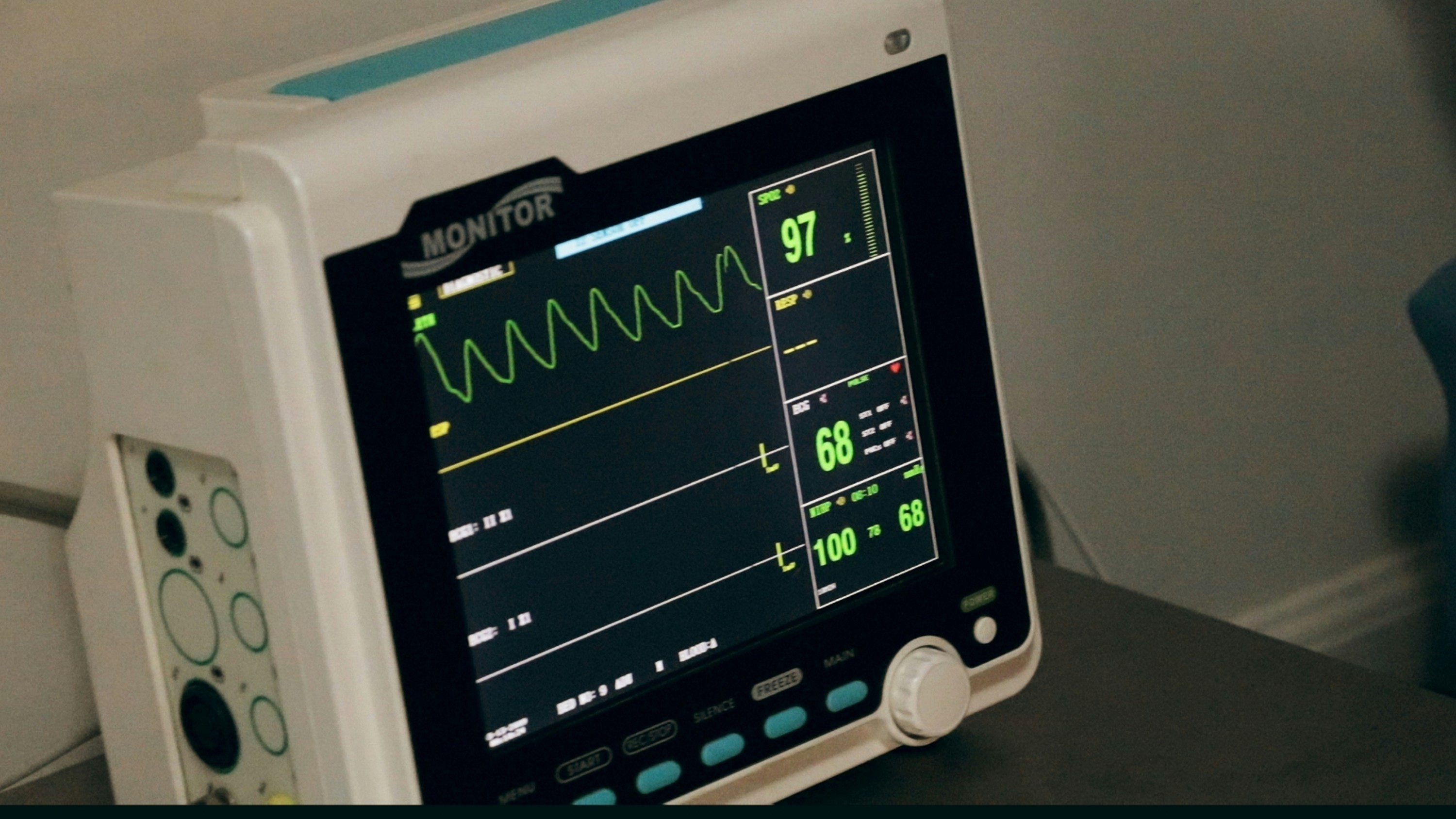 Monitor médico branco com tela preta exibindo gráficos de ondas verdes e números vitais, como 97, 68 e 100, em um ambiente hospitalar