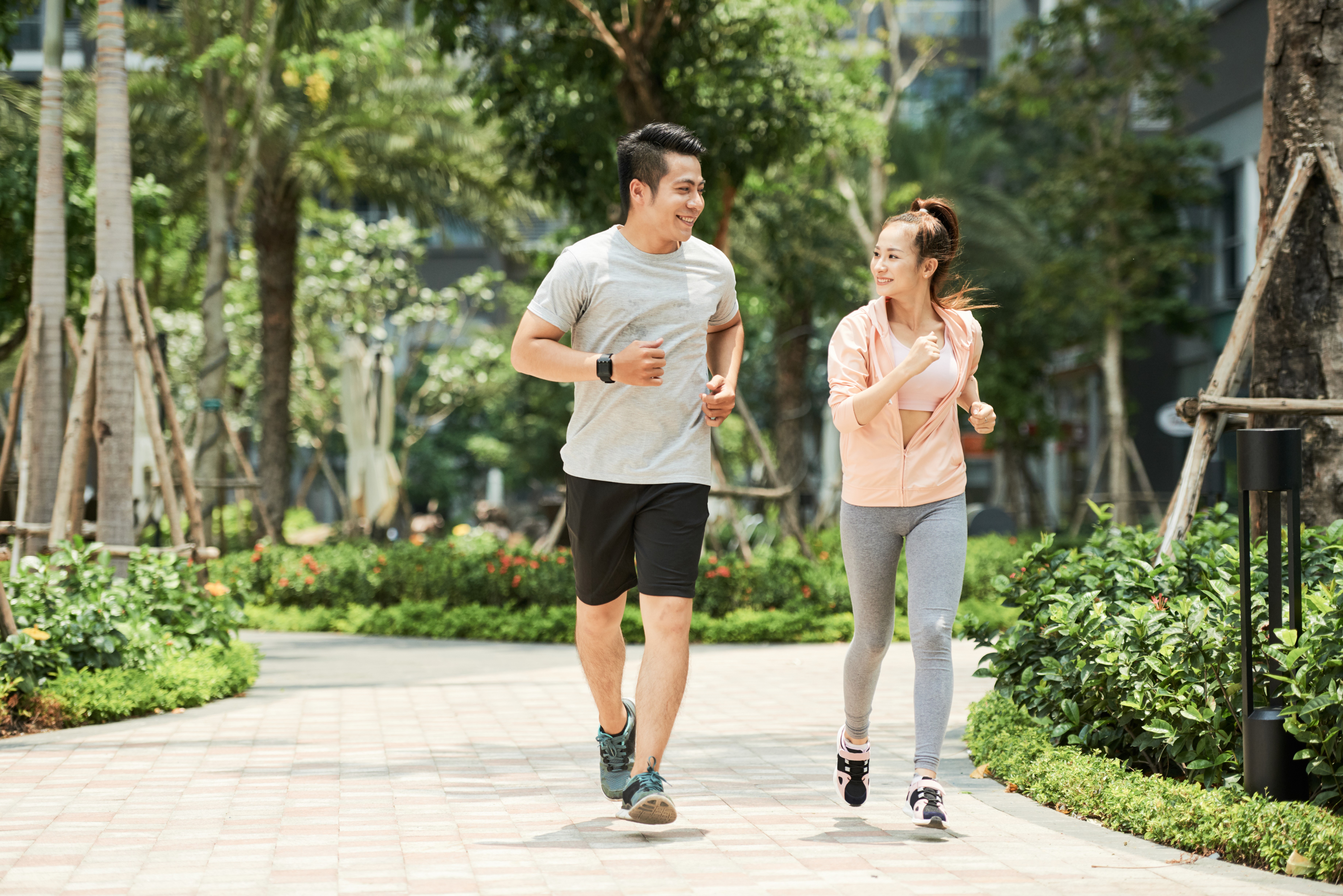 Casal asiático sorridente correndo em um parque arborizado. O homem veste camiseta cinza e shorts pretos, a mulher usa top rosa, jaqueta pêssego e calça cinza. Ambos olham um para o outro, com expressão feliz, enquanto se exercitam em um dia ensolarado