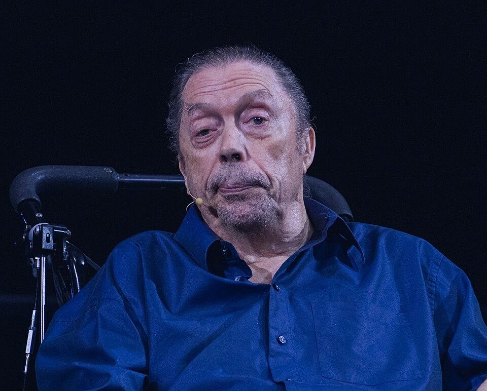 Homem idoso, com cabelo grisalho preso e barba rala, sentado em cadeira de rodas, vestindo camisa azul-marinho e calça jeans escura. Ele tem um microfone preso à gola da camisa e olha para frente com expressão séria. O fundo é escuro