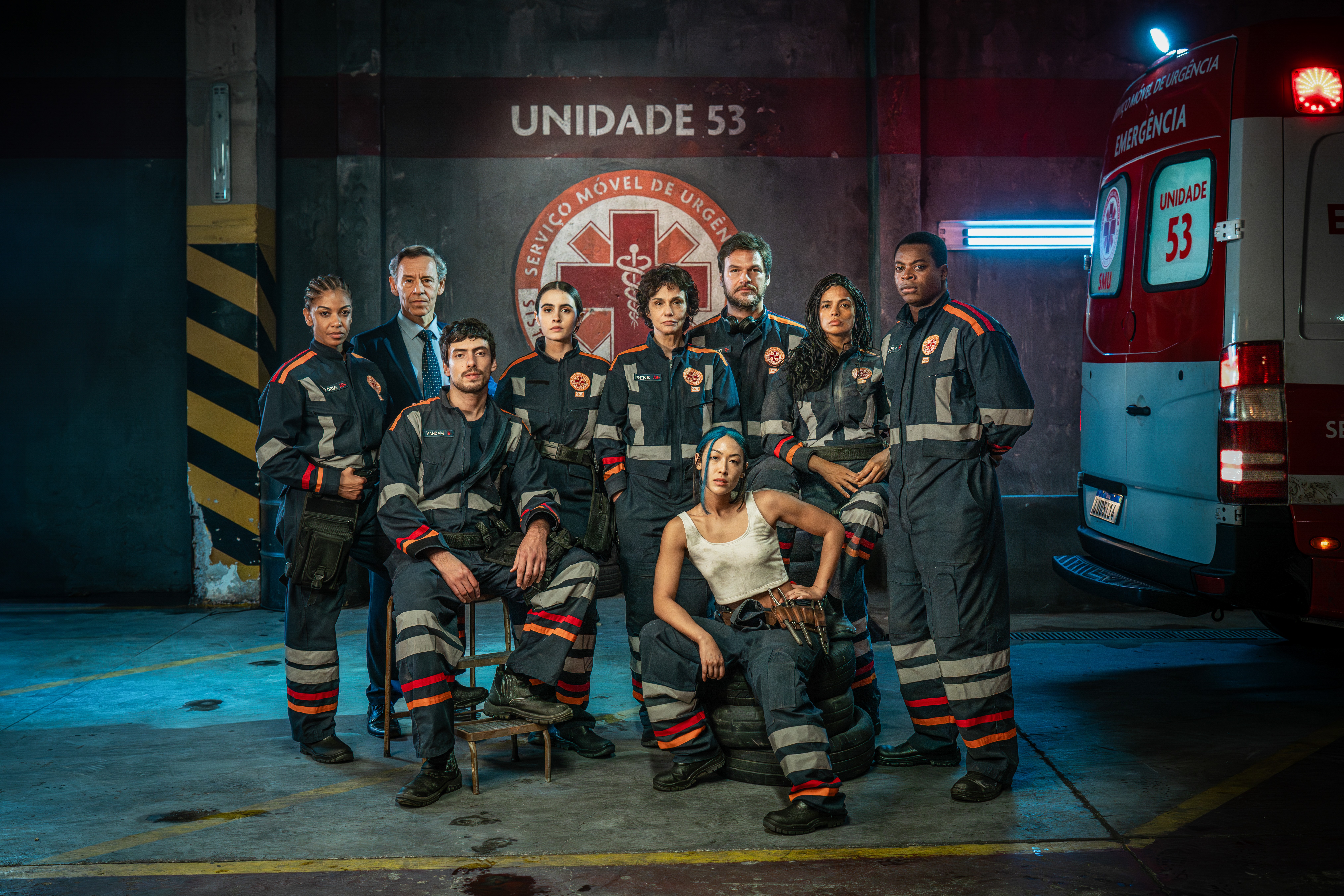 Elenco de série, nove pessoas, em um galpão escuro com uma ambulância vermelha e branca à direita. Oito usam uniformes azuis com faixas refletivas laranja e cinza, e um homem de terno. Uma mulher sentada na frente usa regata branca e calça azul. Ao fundo, um logo vermelho com uma serpente e um bastão, e o texto UNIDADE 53 SERVIÇO MÓVEL DE URGÊNCIA