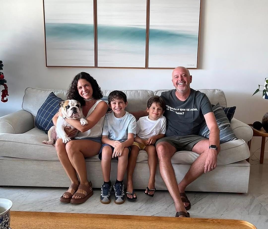 Família sorridente sentada em um sofá claro. A mulher de cabelo cacheado segura um buldogue branco e marrom. Dois meninos, um com camiseta azul e outro com camiseta branca, sentam ao lado dela. O homem careca, com camiseta escura e barba, senta na ponta. Ao fundo, três quadros com ondas do mar e uma decoração natalina discreta