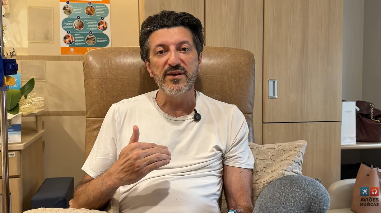 Homem de meia-idade, barba e cabelo escuros, camiseta clara, sentado em poltrona marrom, gesticula com a mão direita. Ele tem um microfone preso à camisa e um acesso venoso no braço esquerdo. Ao fundo, um armário de madeira clara e um pôster informativo na parede. Uma bolsa marrom e uma sacola Aviões Músicas estão à direita